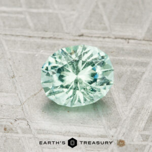 The 0.83-Carat Maine Tourmaline, a round faceted light green gemstone, rests on a textured metallic surface with the Earth's Treasury logo below.