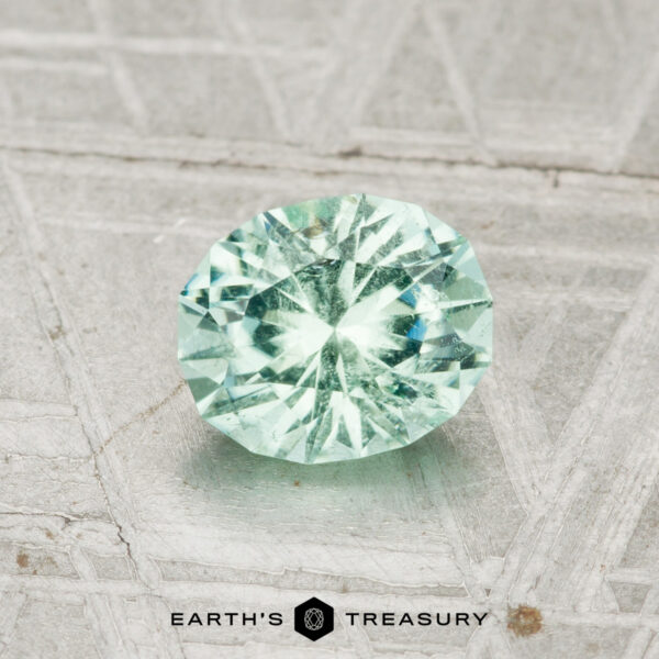 The 0.83-Carat Maine Tourmaline, a round faceted light green gemstone, rests on a textured metallic surface with the Earth's Treasury logo below.