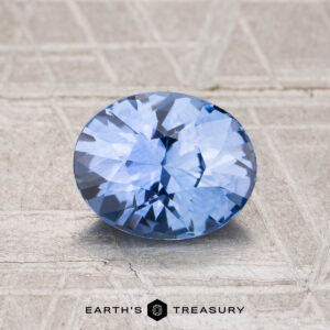 A 1.54-carat heated Ceylon sapphire, oval-shaped and blue, sits on a textured light gray surface with "Earth's Treasury" displayed at the bottom.