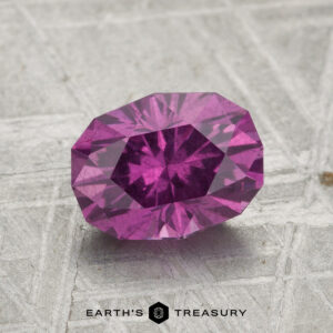 A 1.83-carat pastel pyrope garnet is displayed on a textured gray surface with "EARTH'S TREASURY" at the bottom.