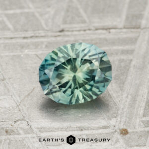 A 1.14-carat Montana sapphire gemstone, faceted in an oval shape and teal in color, is displayed on a textured gray surface with the Earth's Treasury logo beneath it.