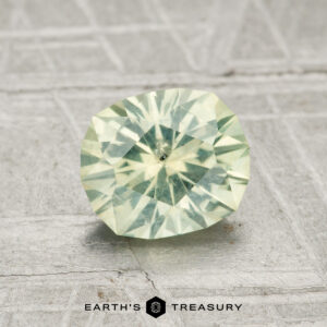 A 1.35-carat round brilliant-cut Montana sapphire with a light green hue rests on a textured gray surface, accompanied by the text "EARTH'S TREASURY" below.