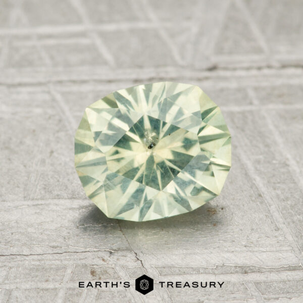 1.35-Carat Light Yellowish Green Montana Sapphire A 1.35-carat round brilliant-cut Montana sapphire with a light green hue rests on a textured gray surface, accompanied by the text "EARTH'S TREASURY" below.