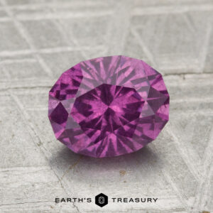 A 1.75-carat pastel pyrope garnet, cut in a faceted oval shape, rests on a textured gray surface with "EARTH'S TREASURY" at the bottom.