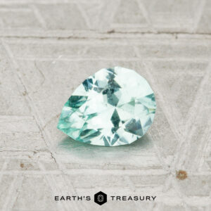 A 0.57-carat Maine Tourmaline, cut in a pear shape with faceted pale green hues, rests on a textured gray surface. The words "EARTH'S TREASURY" appear at the bottom.