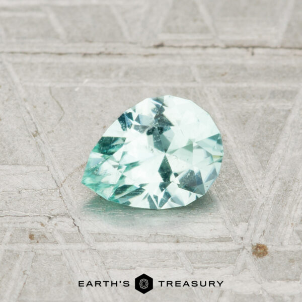 0.57-Carat Maine Tourmaline A 0.57-carat Maine Tourmaline, cut in a pear shape with faceted pale green hues, rests on a textured gray surface. The words "EARTH'S TREASURY" appear at the bottom.