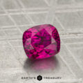 A cushion-cut, vivid pink 1.41-carat ruby (heated) rests on a textured metallic surface, with the "EARTH'S TREASURY" logo and text displayed at the bottom.