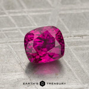 A cushion-cut, vivid pink 1.41-carat ruby (heated) rests on a textured metallic surface, with the "EARTH'S TREASURY" logo and text displayed at the bottom.