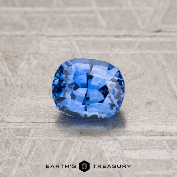 0.98-Carat Ceylon Sapphire (Heated) A 0.98-carat heated Ceylon sapphire, cushion-cut and blue, rests on a textured gray surface featuring the "EARTH'S TREASURY" logo at the bottom.