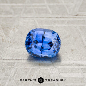 A 0.98-carat heated Ceylon sapphire, cushion-cut and blue, rests on a textured gray surface featuring the "EARTH'S TREASURY" logo at the bottom.