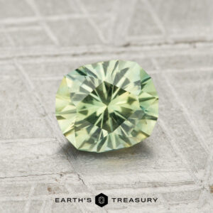 A 1.15-carat Montana Sapphire with a pale green faceted cut is displayed on a textured grey surface, featuring the text "EARTH'S TREASURY" at the bottom.