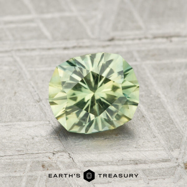 1.15-Carat Green-Yellow Particolored Montana Sapphire A 1.15-carat Montana Sapphire with a pale green faceted cut is displayed on a textured grey surface, featuring the text "EARTH'S TREASURY" at the bottom.