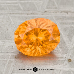 A 2.67-carat Spessartine Garnet gemstone is shown on a textured gray surface with "EARTH'S TREASURY" visible at the bottom.