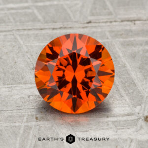 A 1.96-carat round, faceted Spessartine Garnet gemstone is displayed on a textured gray surface with "EARTH'S TREASURY" at the bottom.