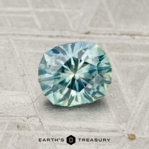 A faceted, oval-cut 1.15-carat Montana sapphire in blue-green tones rests on a textured light gray surface with "EARTH'S TREASURY" beneath it.