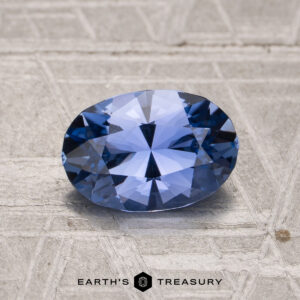 A 1.56-carat Ceylon sapphire, cut in a faceted oval shape, rests on a textured gray surface with the text "EARTH'S TREASURY" below.