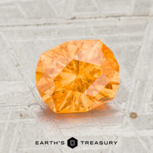 A faceted 1.96-Carat Spessartine Garnet gemstone is showcased on a textured gray surface, with the text "Earth's Treasury" and a geometric logo positioned beneath the stone.