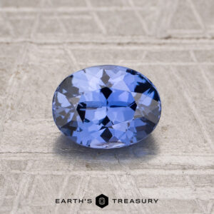 A 1.40-carat Ceylon sapphire, cut in a faceted oval shape, is shown on a textured gray surface with "Earth's Treasury" below.