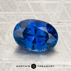 2.42-Carat Ceylon Sapphire (Heated) in oval cut shown on a textured gray surface, featuring the "Earth's Treasury" logo and text at the bottom.