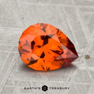 A 1.95-carat pear-shaped faceted Spessartine Garnet with an orange hue rests on a textured gray surface, accompanied by the text "Earth's Treasury" at the bottom.