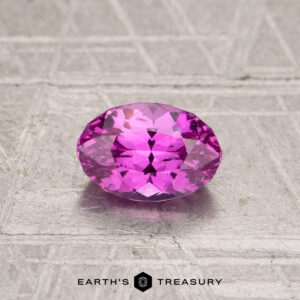 A 1.08-carat oval-cut pink sapphire with multiple facets rests on a textured, light-colored surface. The text "EARTH'S TREASURY" appears at the bottom.