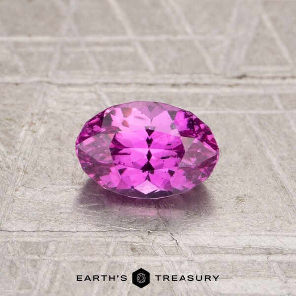 1.08-Carat Sapphire A 1.08-carat oval-cut pink sapphire with multiple facets rests on a textured, light-colored surface. The text "EARTH'S TREASURY" appears at the bottom.
