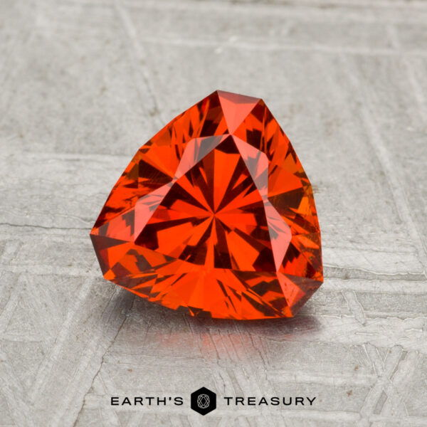 1.69-Carat Spessartine Garnet A 1.69-carat faceted triangular Spessartine Garnet is displayed on a light gray textured surface with the text "Earth's Treasury" below it.