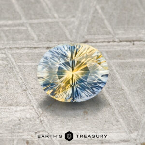 A 0.91-carat faceted Ceylon sapphire (heated), displaying blue and yellow hues, rests on a textured gray surface with "EARTH'S TREASURY" at the bottom.