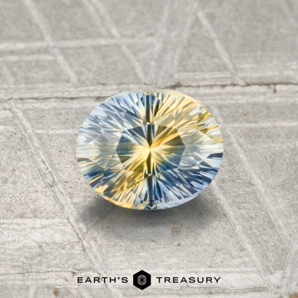 0.91-Carat Ceylon Sapphire (Heated) A 0.91-carat faceted Ceylon sapphire (heated), displaying blue and yellow hues, rests on a textured gray surface with "EARTH'S TREASURY" at the bottom.