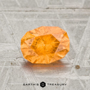 A 1.66-carat Spessartine Garnet with orange facets sits on a textured grey surface, displaying the "EARTH'S TREASURY" logo below.