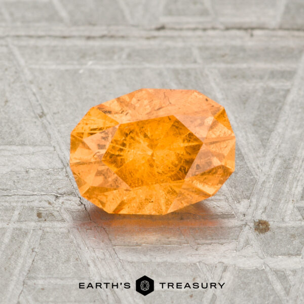 1.66-Carat Spessartine Garnet A 1.66-carat Spessartine Garnet with orange facets sits on a textured grey surface, displaying the "EARTH'S TREASURY" logo below.