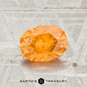 A 1.66-carat Spessartine Garnet with orange facets sits on a textured grey surface, displaying the "EARTH'S TREASURY" logo below.