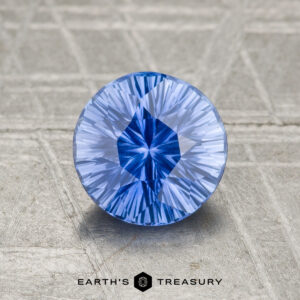 A round, faceted 1.21-carat Ceylon Sapphire (Heated) in vivid blue rests on a textured gray surface, with "EARTH'S TREASURY" and a hexagon logo displayed below it.