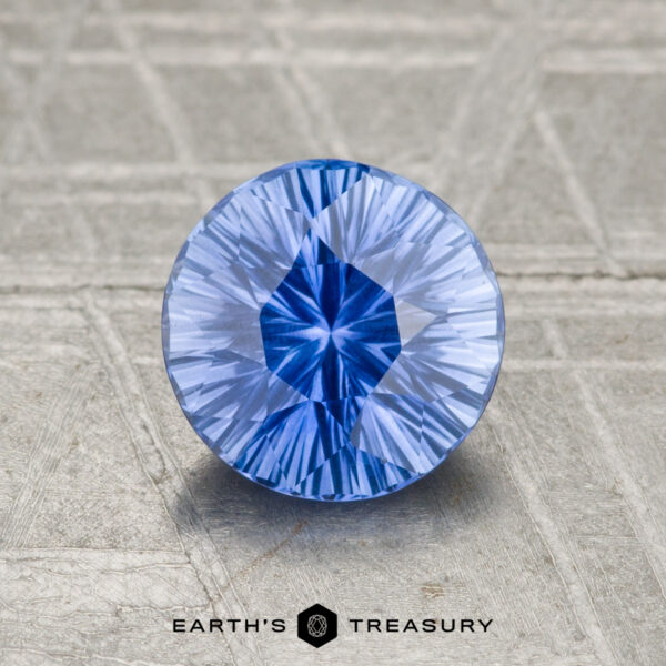 A round, faceted 1.21-carat Ceylon Sapphire (Heated) in vivid blue rests on a textured gray surface, with "EARTH'S TREASURY" and a hexagon logo displayed below it.