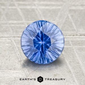 A round, faceted 1.21-carat Ceylon Sapphire (Heated) in vivid blue rests on a textured gray surface, with "EARTH'S TREASURY" and a hexagon logo displayed below it.