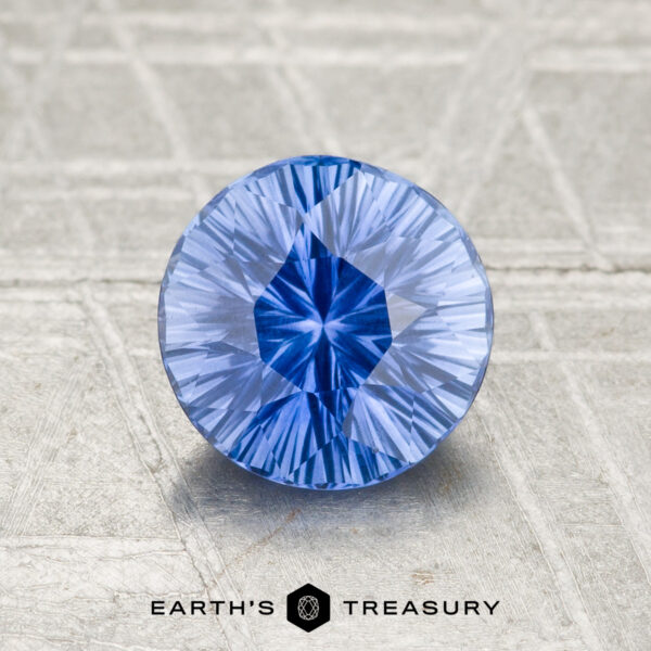 A round, faceted 1.21-carat Ceylon Sapphire (Heated) in vivid blue rests on a textured gray surface, with "EARTH'S TREASURY" and a hexagon logo displayed below it.