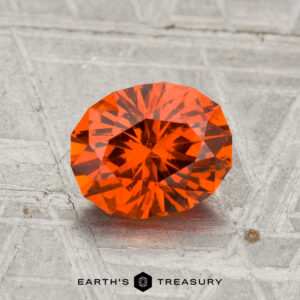 A 1.65-carat Spessartine Garnet, faceted in an oval shape with a vibrant orange hue, is shown on a textured metallic surface with "EARTH'S TREASURY" at the bottom.