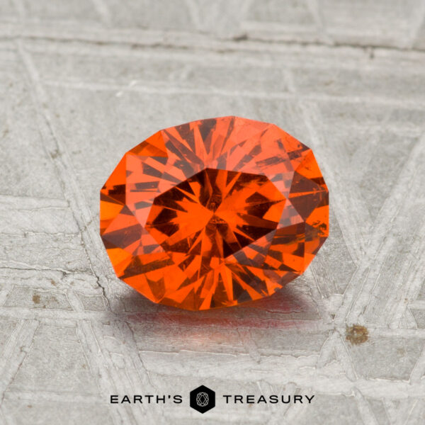 A 1.65-carat Spessartine Garnet, faceted in an oval shape with a vibrant orange hue, is shown on a textured metallic surface with "EARTH'S TREASURY" at the bottom.