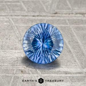 A round, precision-cut 1.02-carat Ceylon Sapphire (heated) with a vivid blue hue rests on a textured metallic surface, featuring the Earth's Treasury logo at the bottom.