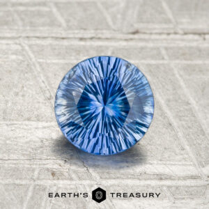 A round, precision-cut 1.02-carat Ceylon Sapphire (heated) with a vivid blue hue rests on a textured metallic surface, featuring the Earth's Treasury logo at the bottom.