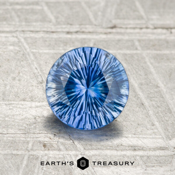 A round, precision-cut 1.02-carat Ceylon Sapphire (heated) with a vivid blue hue rests on a textured metallic surface, featuring the Earth's Treasury logo at the bottom.