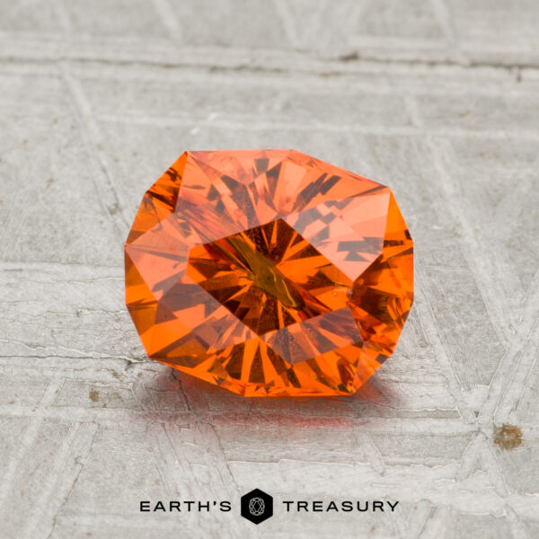 1.58-Carat Spessartine Garnet A 1.58-carat Spessartine Garnet with sharp facets and vivid orange color rests on a textured gray surface, with "Earth's Treasury" displayed below.