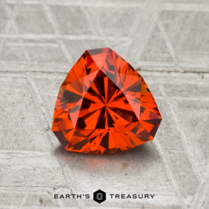 A 1.48-carat spessartine garnet with a faceted triangular cut is displayed on a textured gray surface, featuring the words "EARTH'S TREASURY" at the bottom.