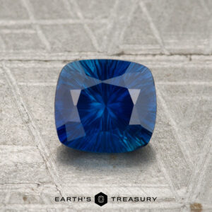 A 1.17-carat heated Ceylon sapphire with a cushion cut is displayed on a textured metallic surface, featuring the "EARTH'S TREASURY" logo and text at the bottom.