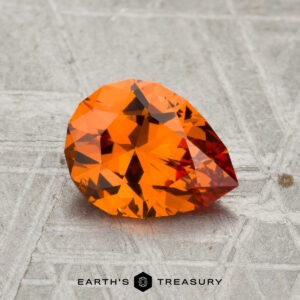 A 1.47-carat faceted pear-shaped Spessartine garnet rests on a textured metallic surface with the text "Earth's Treasury" below.