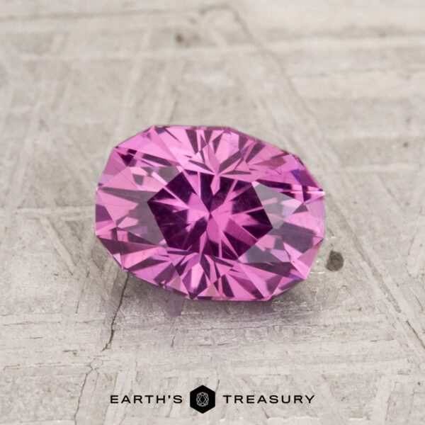 1.38-Carat Pastel Pyrope Garnet A 1.38-carat pastel pyrope garnet gemstone is shown on a gray textured surface, featuring the "EARTH'S TREASURY" logo and text at the bottom.