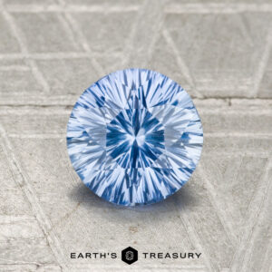 A round, blue faceted 1.05-carat Ceylon Sapphire with a starburst pattern is displayed on a textured metallic surface above the text "EARTH'S TREASURY.