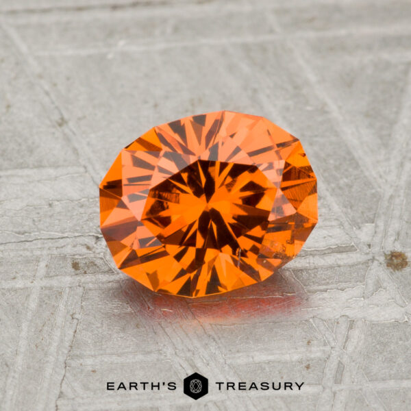 1.39-Carat Spessartine Garnet A 1.39-carat Spessartine Garnet, faceted and round with a vibrant orange hue, is displayed on a textured gray surface bearing the Earth's Treasury logo at the bottom.