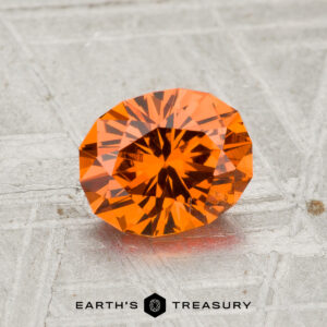 A 1.39-carat Spessartine Garnet with a round brilliant cut is displayed on a textured metallic surface. At the bottom of the image, the text "EARTH'S TREASURY" and a hexagonal logo are visible.