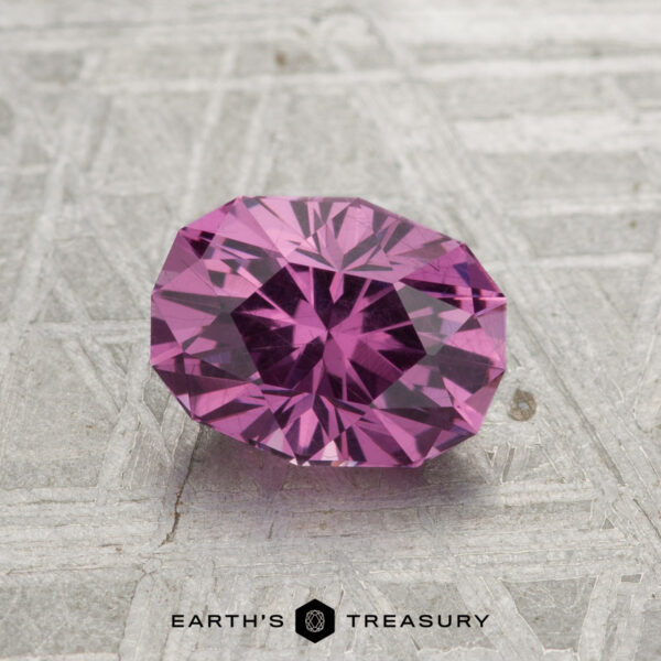 1.38-Carat Pastel Pyrope Garnet A 1.38-carat pastel pyrope garnet, faceted in an oval cut, rests on a textured metallic surface featuring "EARTH'S TREASURY" and its logo at the bottom.